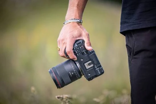 Рука оператора тримає чорну камеру Canon EOS C50 з портретним об'єктивом на рівні стегна, фон розмитий, зелена зона на природі.