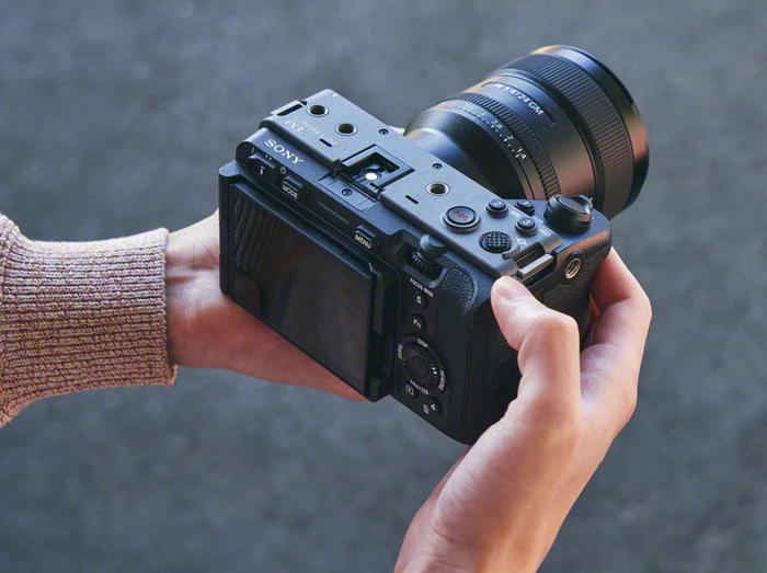 Оператор тримає камеру Sony FX3 з довгим зум-об'єктивом двома руками, знімальний дисплей відкинутий убік, фон текстурований сірий